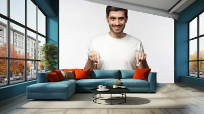 Attractive young man in white t-shirt pointing down with two fingers, isolated on gray background Wall mural