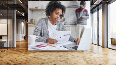 Beautiful young dark-skinned woman keeping hand on touchpad of laptop computer, looking at screen with serious concentrated expression, paying bills online, while her husband making call to bank Wall mural