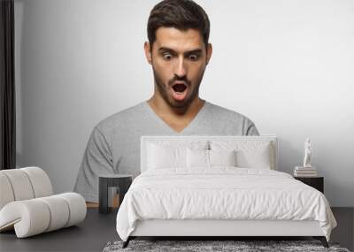 Close up portrait of young man in gray t-shirt, holding smartphone, looking scared and confused reading text message or e-mail Wall mural