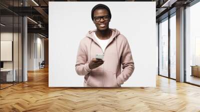 Closeup portrait of young African American man in casual clothes and spectacles pictured isolated on white background holding cellphone in hand looking at camera with open smile, browsing or texting Wall mural