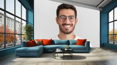 Daylight portrait of young handsome caucasian man isolated on grey background, dressed in white t-shirt and round eyeglasses, looking at camera and smiling positively Wall mural