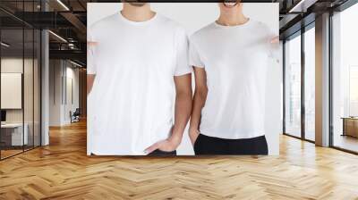 Daylight shot of smiling couple pointing at  blank white t-shirts with index finger, copy space for ads, isolated on gray background Wall mural