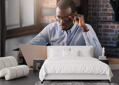 Handsome african business man holding his eyeglasses, working with laptop in modern loft office Wall mural