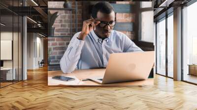 Handsome african businessman holding his eyeglasses, working with laptop in modern loft office Wall mural