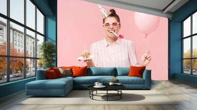 Happy birthday girl wearing pink glasses and holiday hat, holding balloon, cake and whistle in mouth, isolated on studio background Wall mural