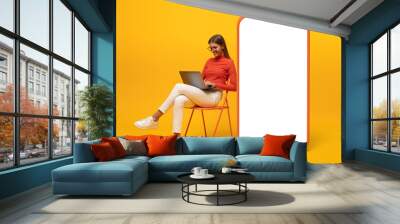 Happy young woman sitting with laptop on chair near blank screen of huge phone, mockup for app Wall mural