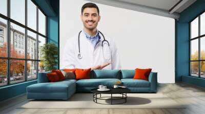 Horizontal banner of smiling young male doctor showing and presenting something with hand, isolated on gray background with copy space on the right side Wall mural