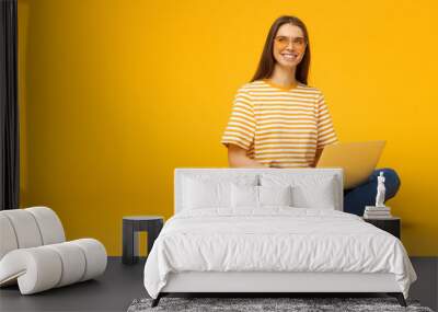 Horizontal banner of young smiling woman sitting on floor holding laptop, isolated on yellow background with copy sapce Wall mural