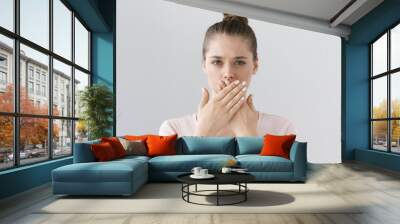 Horizontal photo of young European female isolated on gray background with expression of secrecy and mistrust as she is covering mouth with two hands not willing to disclose something important. Wall mural