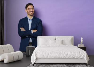 Horizontal shot of young business male standing in jacket and eyeglasses with arms crossed, looking aside with positive smile, isolated on purple background, copyspace on right Wall mural