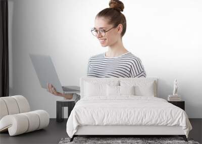 Indoor photo of positive teenage girl standing with laptop, smiling at great content from social networks, pressing fingers to touchpad in order to browse web pages Wall mural