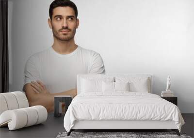 Indoor photo of young European Caucasian man isolated on gray background dressed in white casual T-shirt standing with arms crossed looking upward as if thinking, dreaming or looking for idea Wall mural