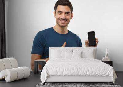 Indoor portrait of attractive young man isolated on grey background, holding blank smartphone, smiling at camera, showing thubms up gesture, feeling happy Wall mural