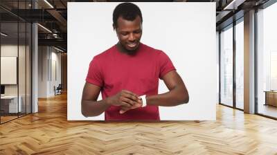 Indoor portrait of handsome African American man standing against grey background in red casual T-shirt adjusting smart wristwatch he is wearing to use it without problems during busy active day Wall mural