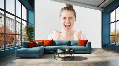 Indoor portrait of young good-looking female, shouting with closed eyes and bumping fists up as if she achieved success and was lucky, looking excited and overemotional, isolated on gray background. Wall mural