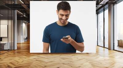 indoor shot of good-looking young male isolated on gray background looking at smartphone, smiling op Wall mural