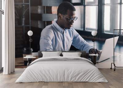 Portrait of young african business man sitting at his desk in loft office, typing message using laptop Wall mural