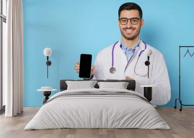 Portrait of young handsome doctor showing phone with blank screen and copy space for medical app ads, isolated on blue background Wall mural