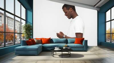 Sideways shot of young attractive african american smiling man looking and at smartphone with while texting his friend, using mobile phone, isolated on gray background Wall mural