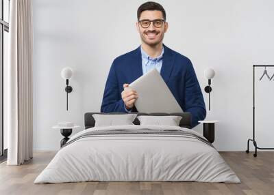 Smiling business man looking at camera through trendy glasses, holding closed laptop next to chest as if going to office, isolated on gray background Wall mural