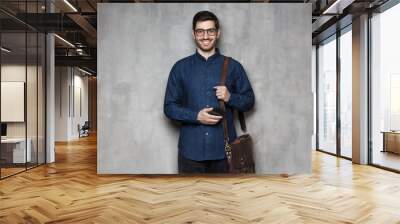 Smiling business man wearing glasses and denim shirt standing against gray textured wall with smart phone in hand and bag on shoulder Wall mural