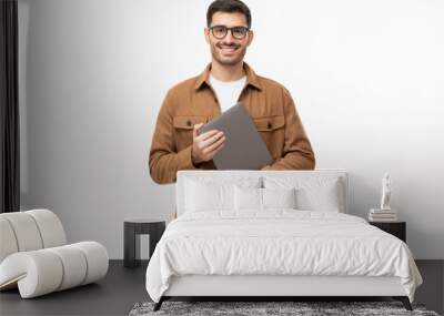 Smiling man looking at camera through trendy glasses, holding closed laptop next to chest, isolated on gray background Wall mural