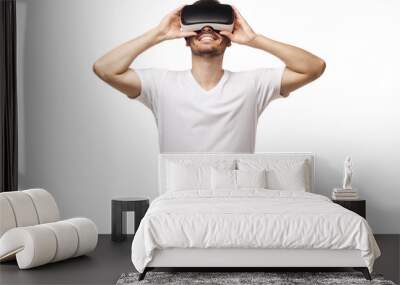 Smiling man wearing tshirt, holding virtual reality headset or VR glasses with both hands, playing video game, isolated on white background Wall mural