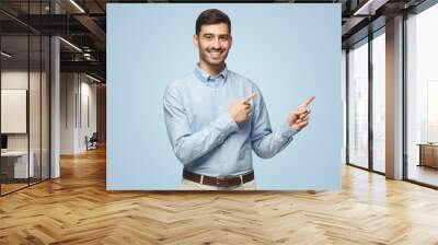 Smiling young business man pointing away, isolated on blue background Wall mural