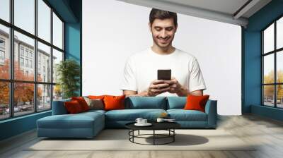 Studio portrait of young european caucasian man isolated on gray background wearing white t-shirt standing in front of camera, looking attentively with smile at screen of smartphone he is holding Wall mural