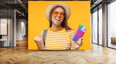 Tourism concept. Excited young female tourist celebrating if she is a winner, holding passport with flight tickets, isolated on yellow background Wall mural