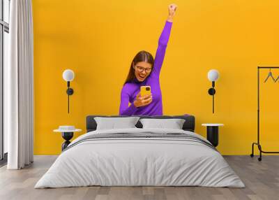 Winner. Excited happy business woman sitting on floor with phone, raising one hand in the air saying yes, isolated on yellow background Wall mural