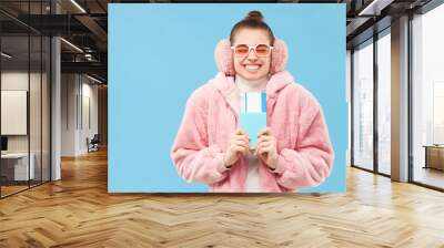 Winter travel. Young woman in pink furry coat holding passport and tickets, feeling happy about travelling, isolated on blue background Wall mural