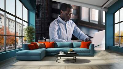 Young african american business man working, using laptop at office while sitting at table with papers and cup of coffee, typing message on computer keyboard Wall mural