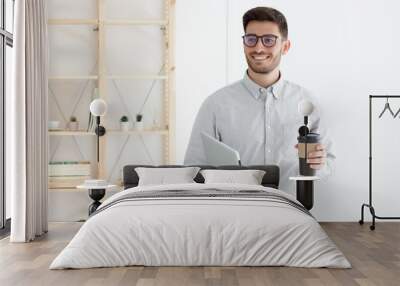 Young business man wearing gray shirt and eyeglasses standing in office, holding laptop, drinking takeaway coffee, looking aside with positive smile Wall mural