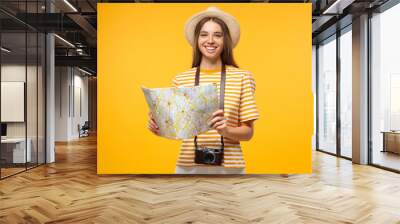 Young cheerful female tourist holding oldschool paper map, isolated on yellow background Wall mural