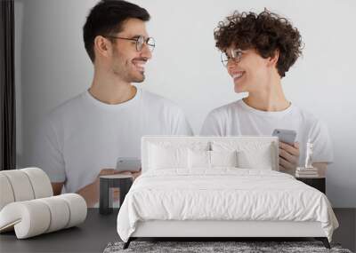 Young couple holding mobile phones, standing together and looking at each other, isolated on gray background Wall mural