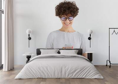 Young female looking at screen of phone with smile, browsing web pages or chatting with friend, standing isolated on gray background Wall mural