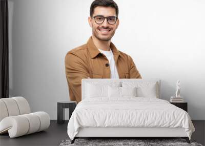 Young hispanic man wearing brown shirt and glasses, looking at camera with positive confident smile, holding arms crossed, isolated on gray background Wall mural