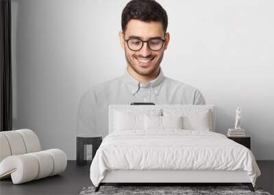 Young man wearing gray shirt and glasses, holding his smart phone and exchanging messages with friends, isolated on studio background Wall mural