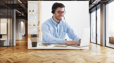 Young man working in hotline answering client inquiries and helping solve problems, feeling positive and confident, smiling happily, typing data on laptop in office Wall mural