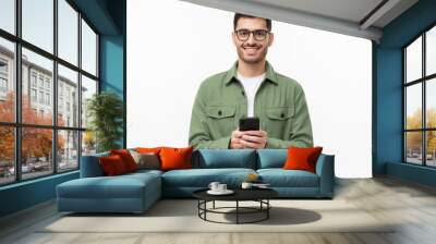 Young modern man in casual green shirt and glasses, holding smart phone in hands, looking at camera with smile, isolated on gray background Wall mural