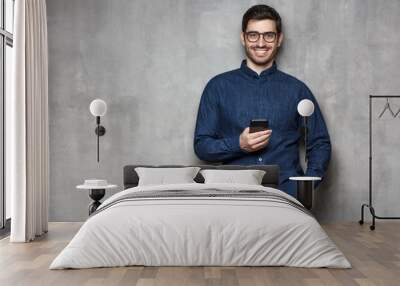 Young smiling man wearing denim shirt and trendy glasses standing against gray wall with cellphone in one hand. Copy space on the left side Wall mural