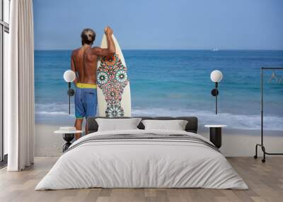 Attractive bearded curly man with a surfboard is standing with his back on a beach and looking to the ocean, located on the left side of the frame Wall mural