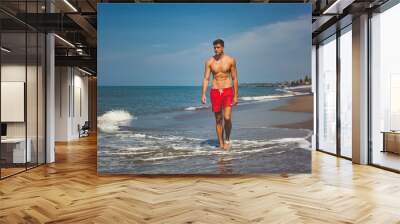 handsome sexy man in red shorts and with naked tanned fit torso walks on beach along the ocean. bright blue water and sky Wall mural