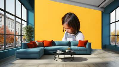 A caucasian girl in a gray dress and white shirt uses and plays with a tablet on a yellow background Wall mural