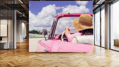 Anonymous female traveler in straw hat driving pink retro cabriolet on cloudy day during summer vacation in Cuba. Unrecognizable woman driving retro cabriolet on city road Wall mural