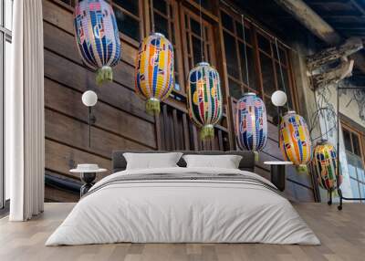 Six colorful paper lanterns hang from the eaves of a wooden building. The lanterns are decorated with geometric patterns and brightly colored stripes in Taiwan. Wall mural