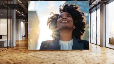 Photo of a happy African American woman with curly hair in a business outfit looking up at city buildings. Wall mural