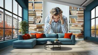 Overwhelmed young adult sitting at a desk surrounded by stacks of books and scattered papers, hands cradling head Wall mural