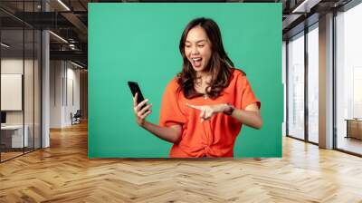 A portrait of an Asian woman wearing an orange shirt, looking surprised while holding a smartphone, isolated on a green background. Wall mural